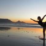 Woman practicing yoga at sunrise on Bloubergstrand beach with Table Mountain in the background.